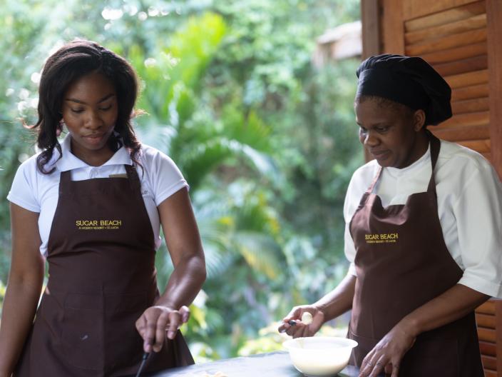 Duas mulheres com aventais castanhos da marca « Sugar Beach » preparam comida numa mesa ao ar livre, cortando ingredientes brancos ao lado de uma tigela.