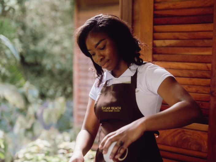 Uma mulher com um avental da « Sugar Beach » deita ingredientes de uma tigela pequena para uma tigela de metal, num balcão ao ar livre.