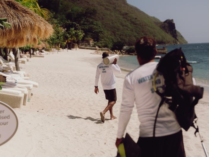 Duas pessoas de camisa branca transportam equipamento de mergulho ao longo de uma praia de areia, onde se avistam guarda-sóis de palha, espreguiçadeiras, água e uma montanha coberta de floresta nas proximidades.