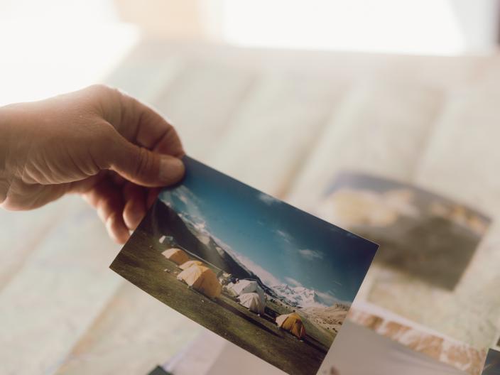 Uma mão segura uma fotografia de tendas ao pé de uma montanha, sobre uma mesa repleta de outras fotografias de viagens e de montanha.