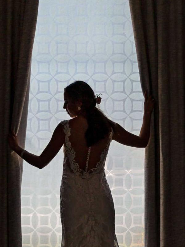 A silhouette of a bride from behind, opening a set of heavy curtains and turning her head to look back towards the camera.
