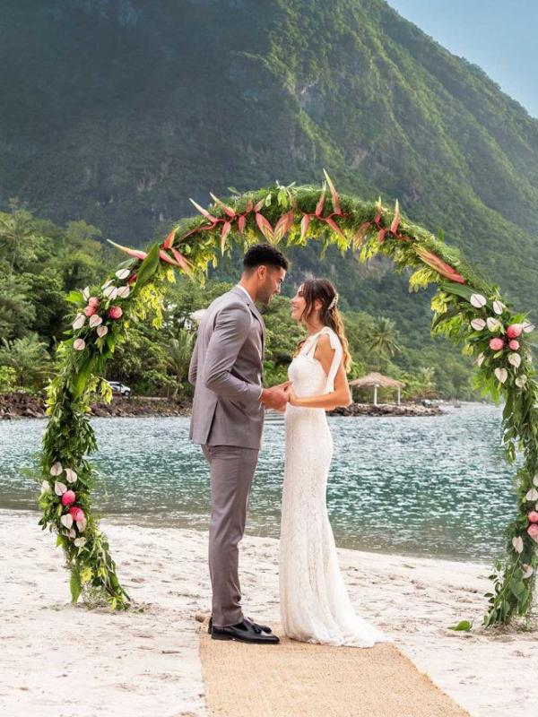 A bride and groom standing under an arch of greenery and flowers while on the beach, with the Pitons in the background.