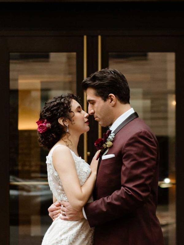 A groom in a burgundy suit with his arms around his bride's waist while they stare lovingly into each other's eyes.
