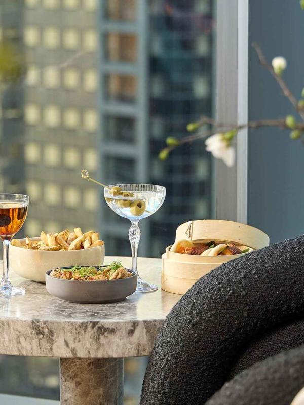 Two cocktails and snacks on a side table at Pandan, with a view of downtown Chicago through a window in the background