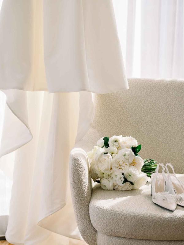 A veil hung up near a white armchair with a bridal bouquet and shoes resting upon it, near a white curtain in a light-streaked hotel room