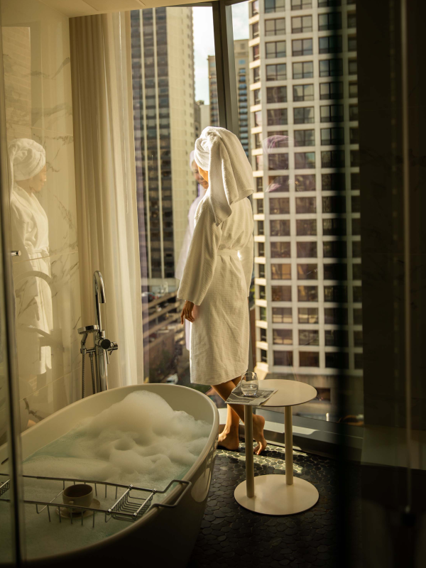 A woman standing in the bathroom in a housecoat and a towel over her hair staring out the window in a room at Viceroy Chicago