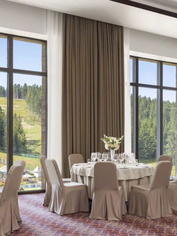 The Aero room at Viceroy Kopaonik Serbia, with padded chairs surrounding round banquet tables on a pink striped carpet, and large windows looking out on a ski slope in summer time