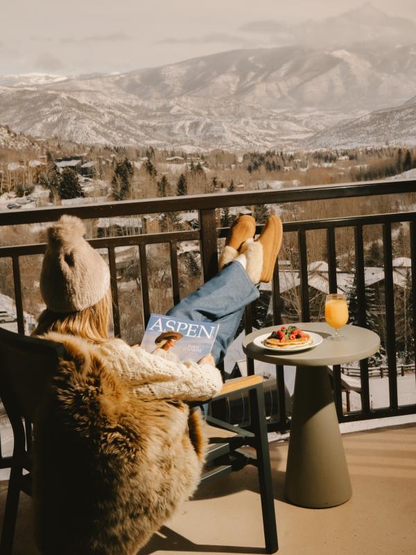 Uma mulher lê uma revista sentada numa cadeira numa varanda com vista para uma cordilheira coberta de neve no Colorado