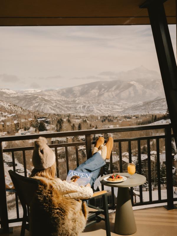 A woman reads a magazine with her feet up, seated in a chair on a balcony overlooking a snowy Colorado mountain range