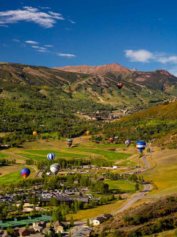 A dozen hot air balloons fly over the town of Snowmass, with the green mountains in the background