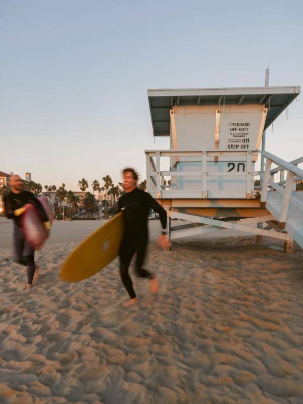 two people in wetsuits carrying longboards run in front of lifeguard station at beach