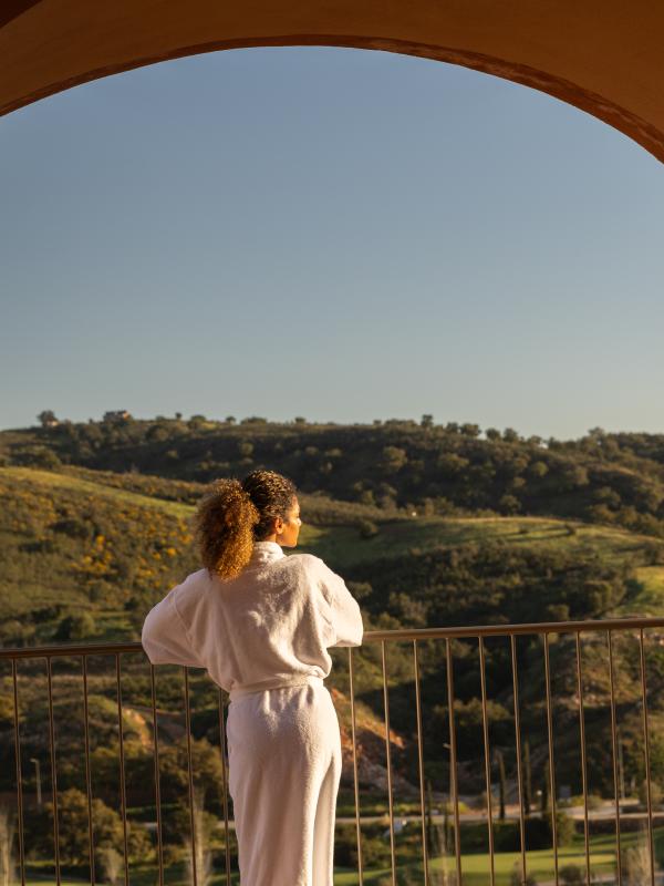 Woman in spa robe looks out over a balcony at rolling green hills.