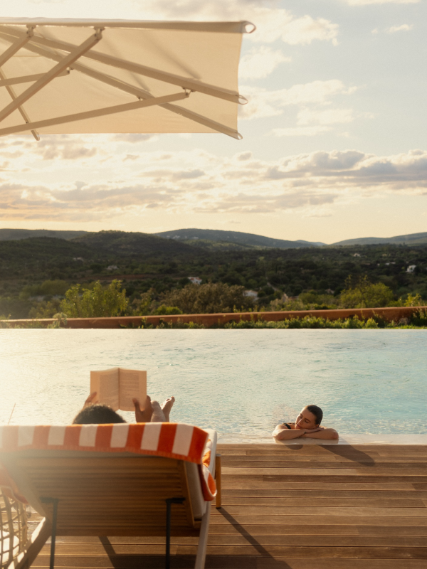 A person sits in a deck chair reading, while their partner lounges in the pool.