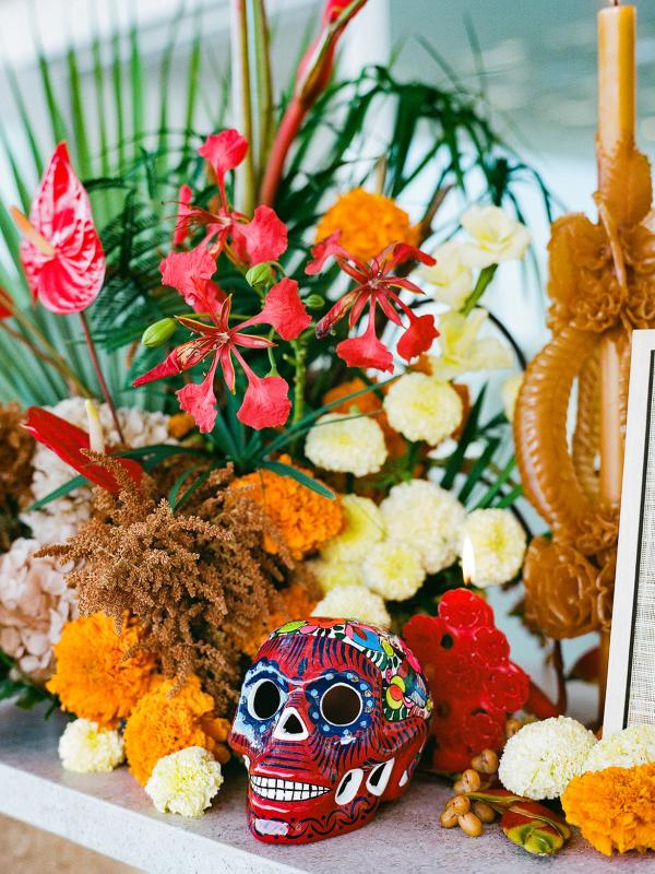 Brightly painted red sugar skull sits before a vibrant Day of the Dead altar arrangement featuring orange marigolds, red flowers, and green foliage next to a tall, molded brown candle.