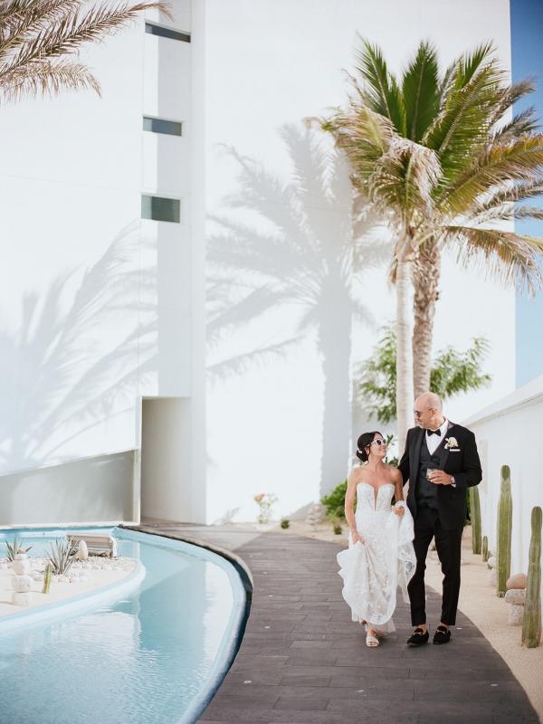 A newly married couple in wedding attire walks along a dark pathway beside a bright blue pool, framed by a white building and tall palm trees casting distinct shadows.