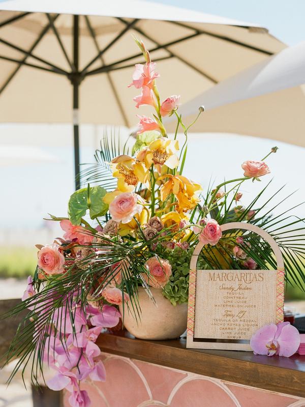 Lush floral arrangement featuring yellow orchids, pink roses, and tropical greenery next to a wooden-framed margarita menu sign on a pink tiled outdoor bar under a large umbrella.