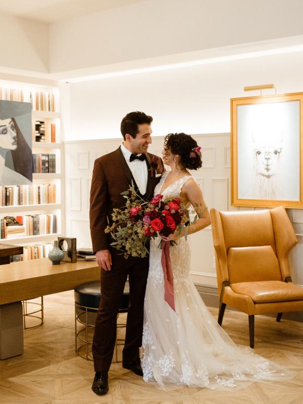 A newly married couple stands close together in a stylish, brightly lit room featuring built-in bookshelves, modern art, and tan leather seating.