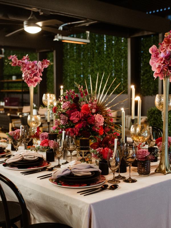 Elegant formal dinner table setting with deep red floral centerpieces, candlelight, and black tableware against a backdrop of a dark patio with green hedging.