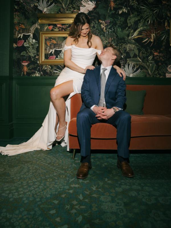 A smiling bride in a white dress sits on the arm of an orange couch looking down at the groom in a blue suit seated below her, set against a dark, tropical-print wallpaper.