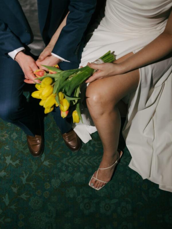 Close-up overhead view of a couple sitting together, holding a bouquet of yellow tulips, with the bride in a white dress and the groom in a blue suit over a dark green patterned rug.