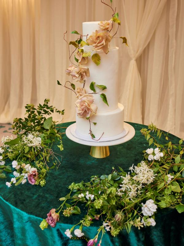 Three-tiered white wedding cake adorned with blush sugar roses and trailing vines, set on a gold stand atop a dark emerald velvet draped table surrounded by lush green and white floral arrangements.