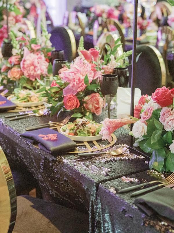 Uma mesa de banquete ricamente decorada apresenta uma toalha preta com lantejoulas, centros de mesa altos com arranjos florais em tons de rosa e vermelho, talheres dourados e guardanapos escuros com bordados coloridos.