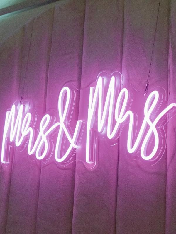 Um letreiro em neon rosa vivo com a inscrição «Mrs & Mrs» ilumina um pano de fundo drapeado, com textura e em tons de rosa-malva.
