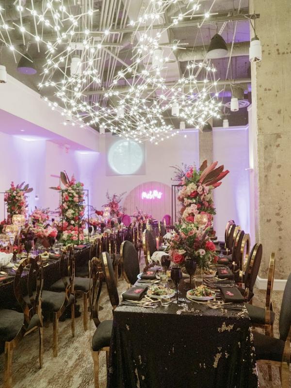 Um espaço para eventos luxuoso, com mesas decoradas com lantejoulas pretas, arranjos florais altos com flores cor-de-rosa e vermelhas, cadeiras com detalhes dourados e uma instalação suspensa espetacular composta por luzes brancas interligadas.
