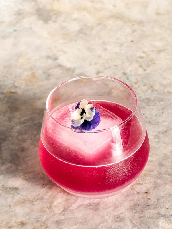 Um cocktail de cor rosa vibrante, servido num copo arredondado, repousa sobre uma superfície de mármore claro, com um grande cubo de gelo decorado com uma pequena flor comestível roxa e branca.