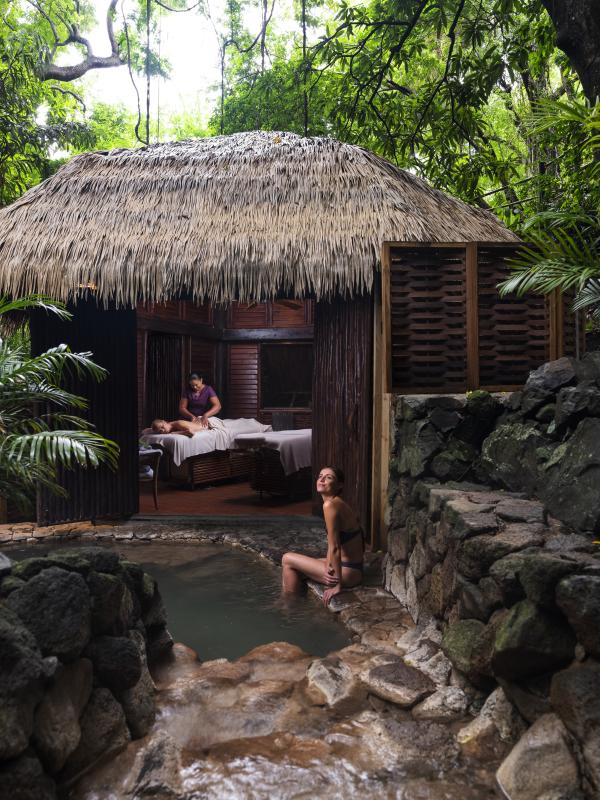 A woman sits at the edge of a natural hot spring next to a thatched-roof spa where a massage is being given.