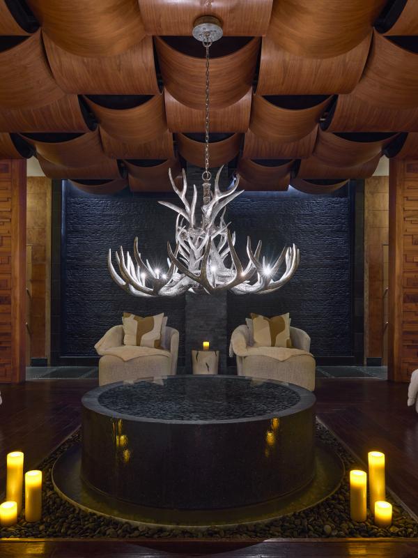 Um salão de spa relaxante com assentos confortáveis, uma fonte central em pedra e um lustre de chifres sob um teto revestido a painéis de madeira.
