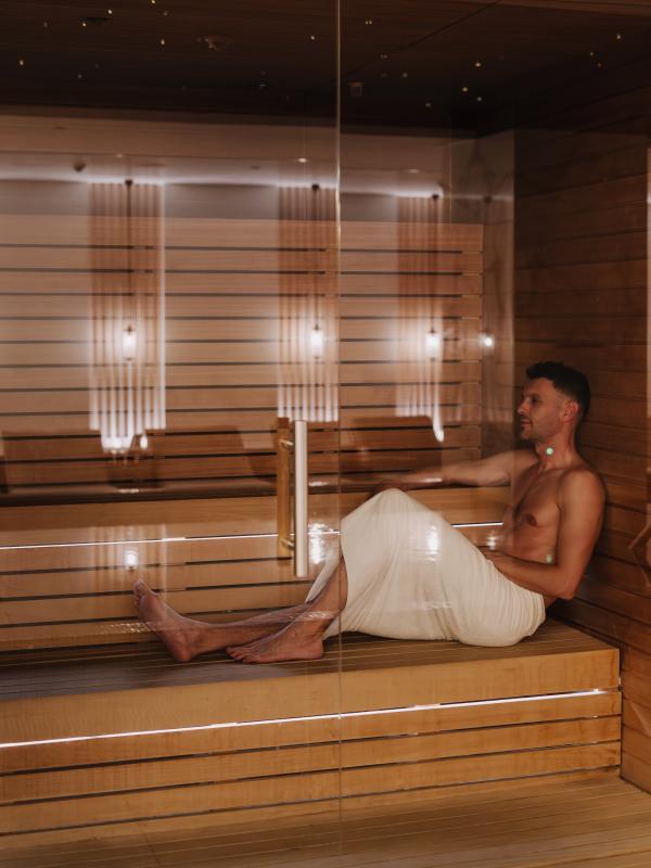 Man in a white towel relaxes on a wooden bench in a modern sauna.