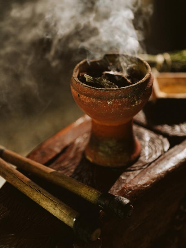 Gros plan sur de l'encens qui se consume dans un bol en terre cuite, accompagné de deux ustensiles en bois, posé sur une surface sombre et texturée.