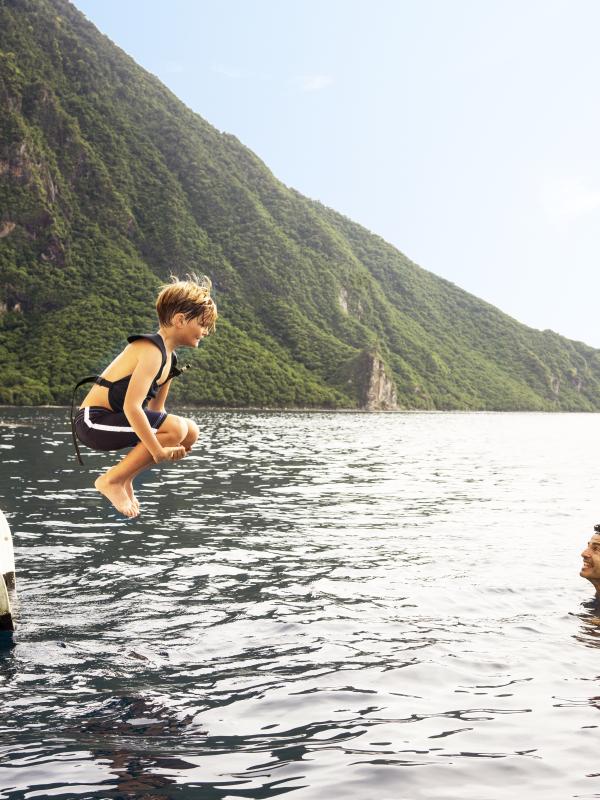 Un garçon en plein saut, en train de plonger depuis un voilier dans l'océan, avec un plongeur en apnée dans l'eau et une montagne verdoyante en arrière-plan.