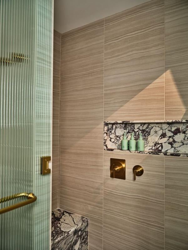 Modern shower with wood-patterned tiles, a marble shelf holding three green bottles, brass fixtures, and a ribbed glass door.
