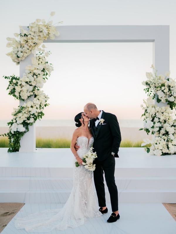 A bride and groom kiss near a square arch decorated with flowers