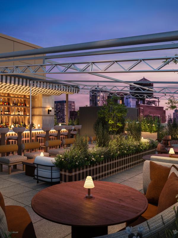 A rooftop restaurant patio at dusk has a bar, cushioned seating, round tables, planters, and a metal canopy overlooking city buildings.