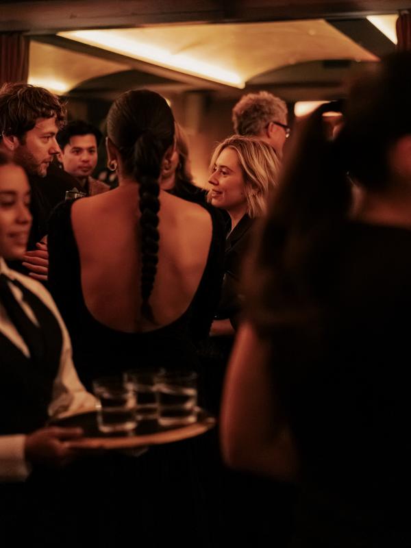 Guests mingle in a warmly lit room; a woman with a long braid and a low-backed black dress faces a blonde woman, while a server carries drinks.