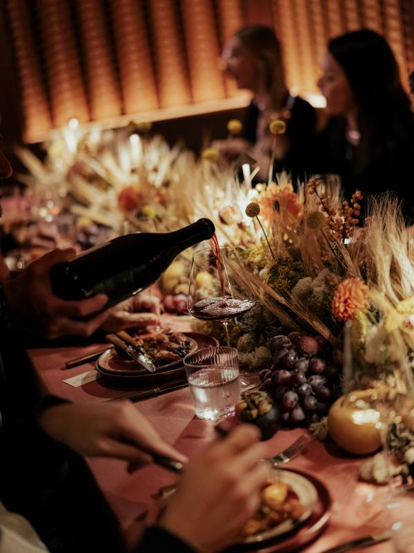 A person pours red wine into a glass at a candlelit dinner table decorated with flowers, fruit, plates, and drinking glasses.
