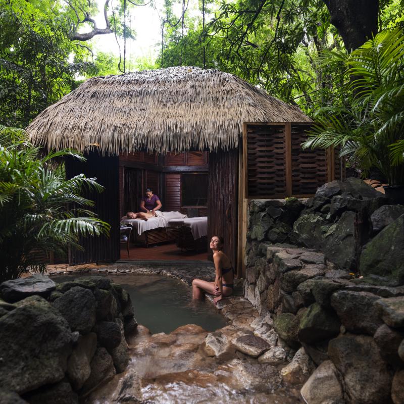 Une femme est assise au bord d'une source thermale naturelle, à côté d'un spa au toit de chaume où l'on procède à un massage.