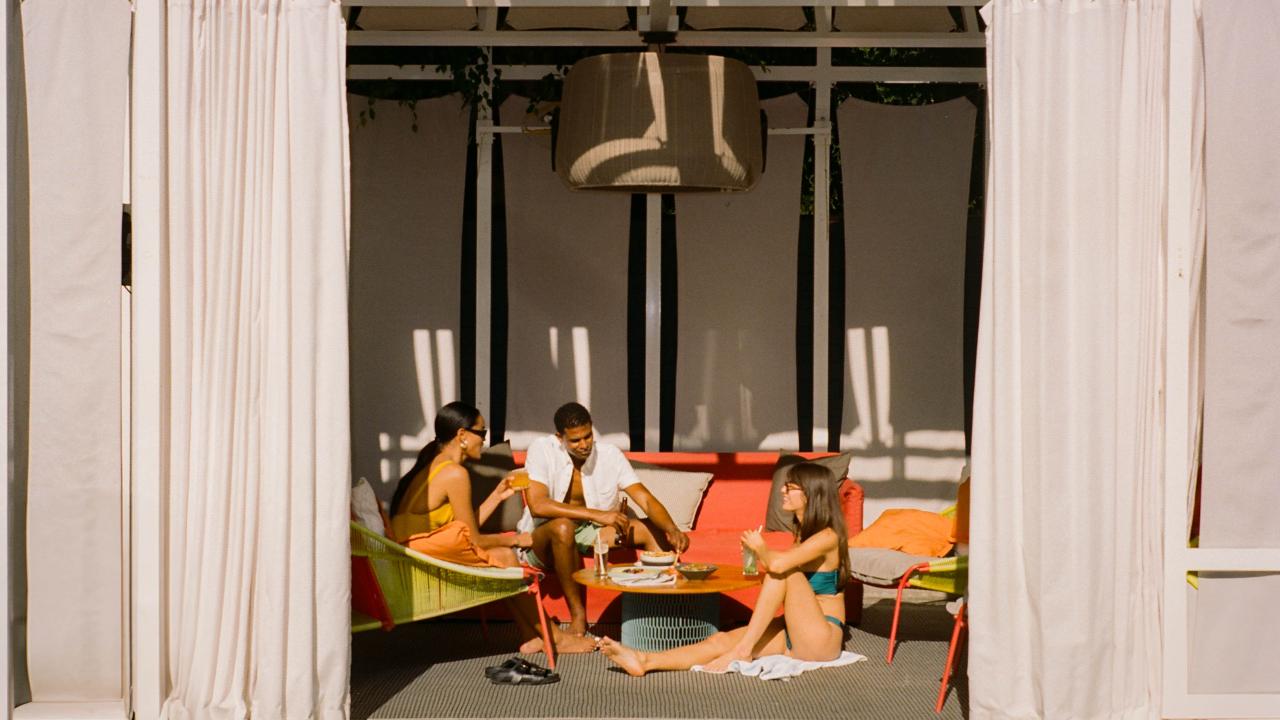 Three friends sit in a cabana, sipping drinks and talking.