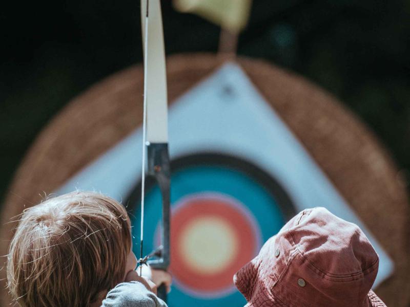 two children holding a bow point at an archery board in the background