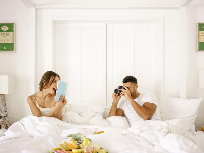 couple sit in bed while holding book and camera