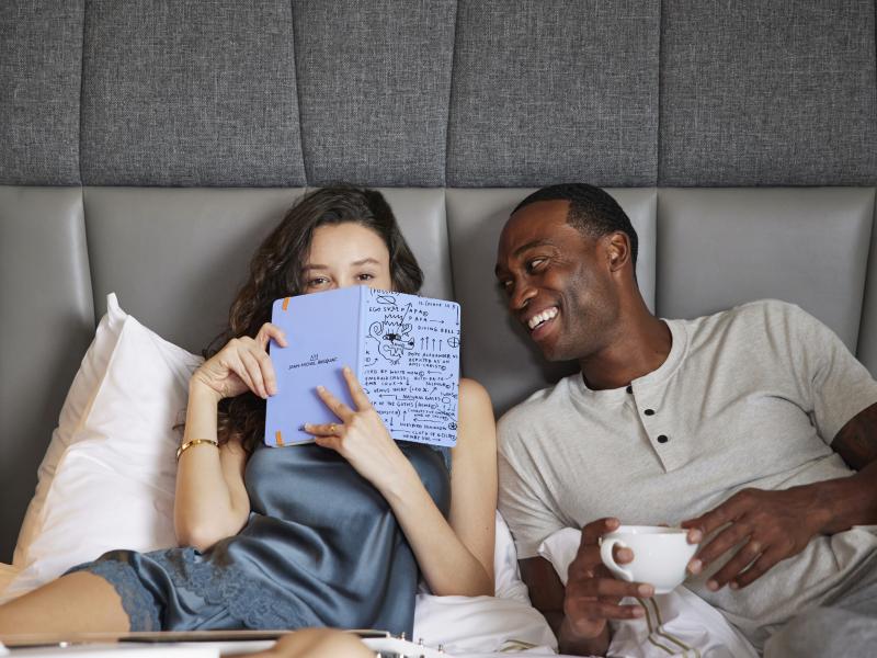 A woman holds a journal up to her face, hiding her smile as she lounges in a bed next to a smiling man holding a coffee cup.