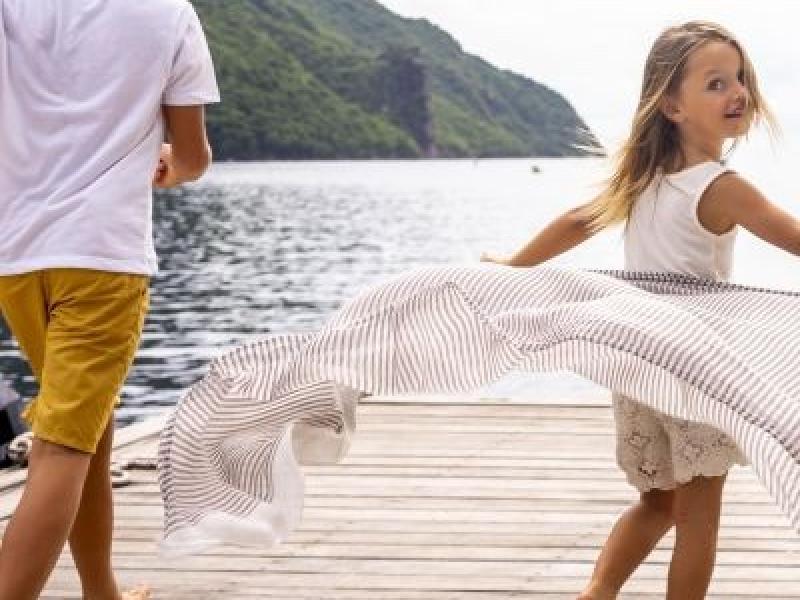 A boy in shorts and a tshirt and a girl in a flowy dress frolic on a dock on a lake at the foot of a mountain.