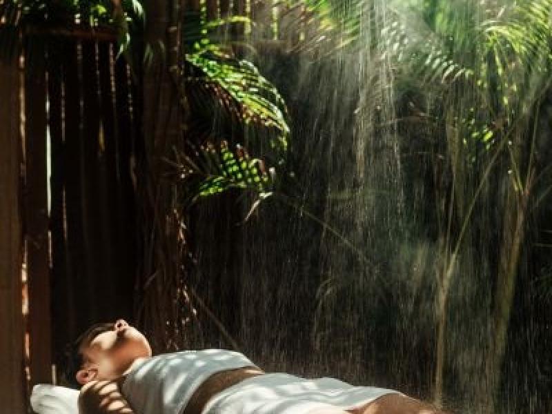 A woman draped in towels lies on an outdoor spa massage table with tropical plants growing around her