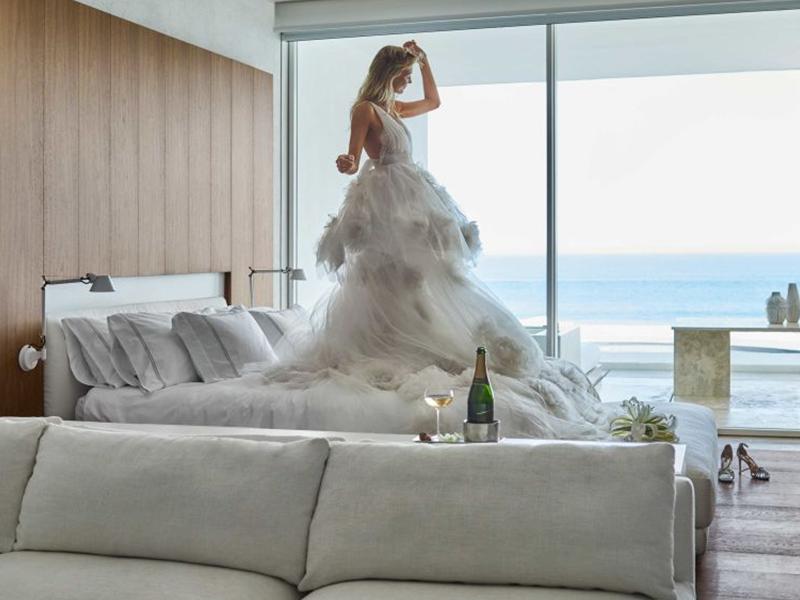 A bride poses in her wedding gown while standing on the bed of an oceanside hotel room