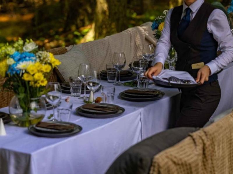 A server sets a table setting at an outdoor dining table with white linens and flowers