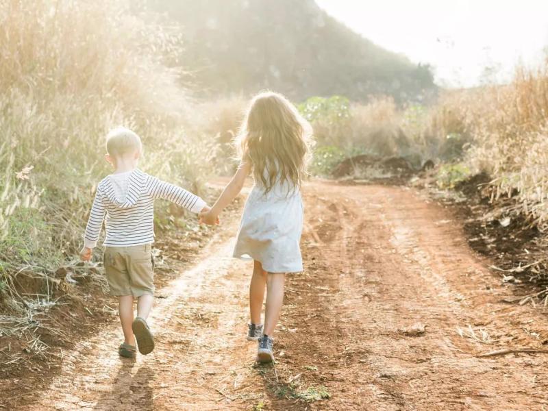 Two children holding hands and walking down a dirt path next to a field on a sunny day.