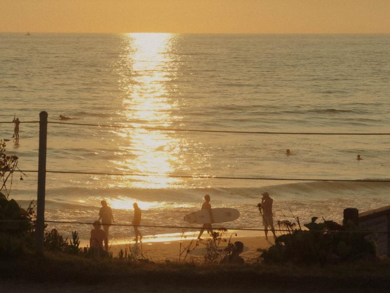 Au coucher du soleil, on aperçoit au bord de la mer les silhouettes de baigneurs et d'un surfeur portant sa planche, tandis que le soleil se reflète avec éclat sur l'eau de la mer.